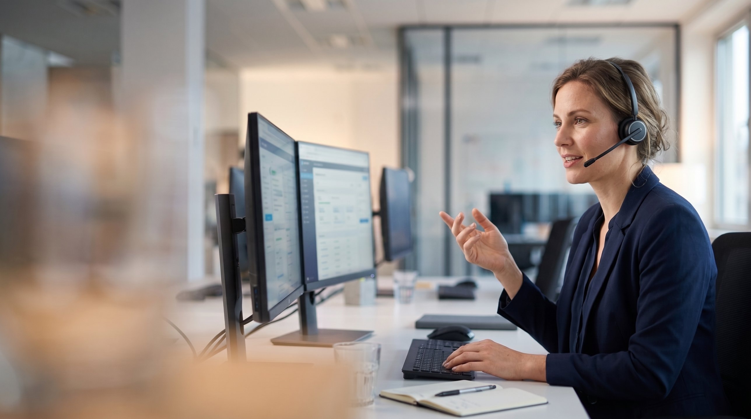 A human call center agent on the phone, several other agents visible working at desks behind them