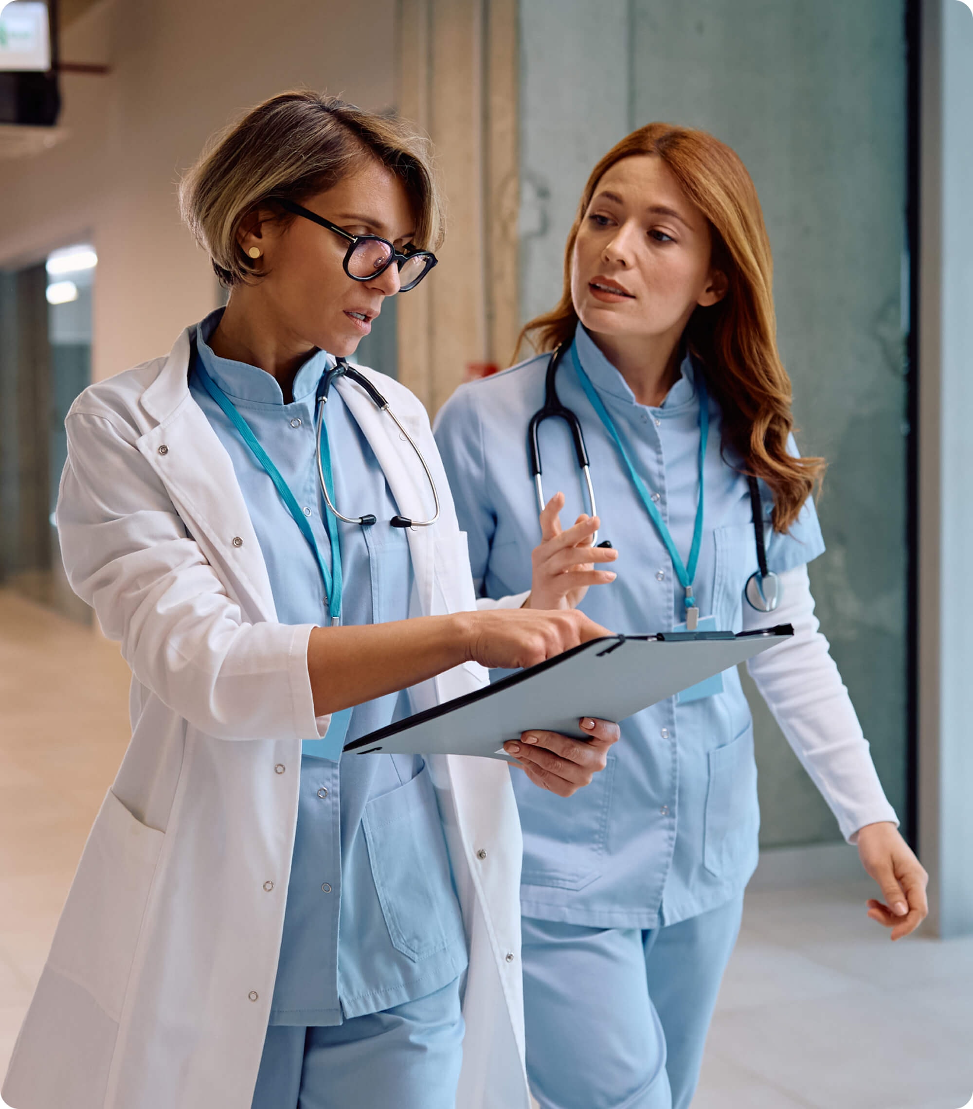 Two healthcare workers in scrubs and a white coat walking down a hospital corridor, reviewing a clipboard